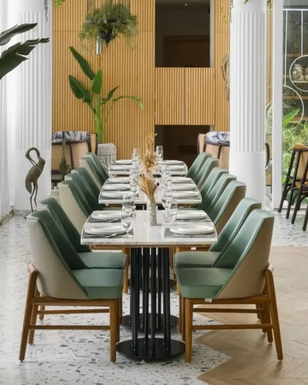 The main dining area is bright and airy, with tall plants and light wood paneling creating a garden-like feel. The arrangement of tables encourages both intimate and group dining, set against a backdrop of serene greenery.