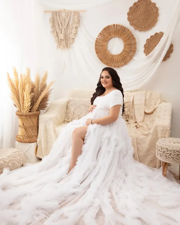 A beautiful full-length portrait in the boho setup. The mother-to-be is seated on the couch, with the long, ruffled train of the white gown cascading around her.