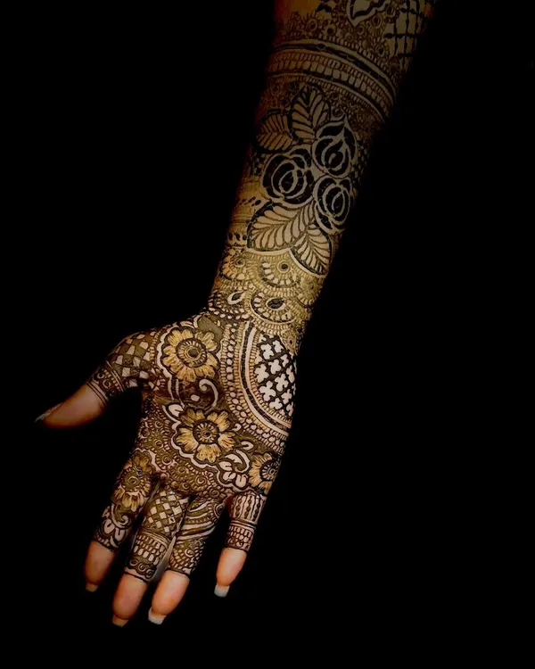 A stunning, professional shot of a full-arm bridal mehendi stain. The dark, rich color and intricate floral details are captured beautifully against the black background.