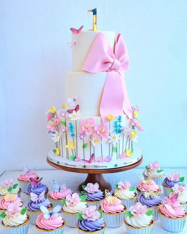 This first birthday dessert spread features a three-tiered floral cake with a large fondant bow, surrounded by matching cupcakes with sugar flowers and butterfly toppers.