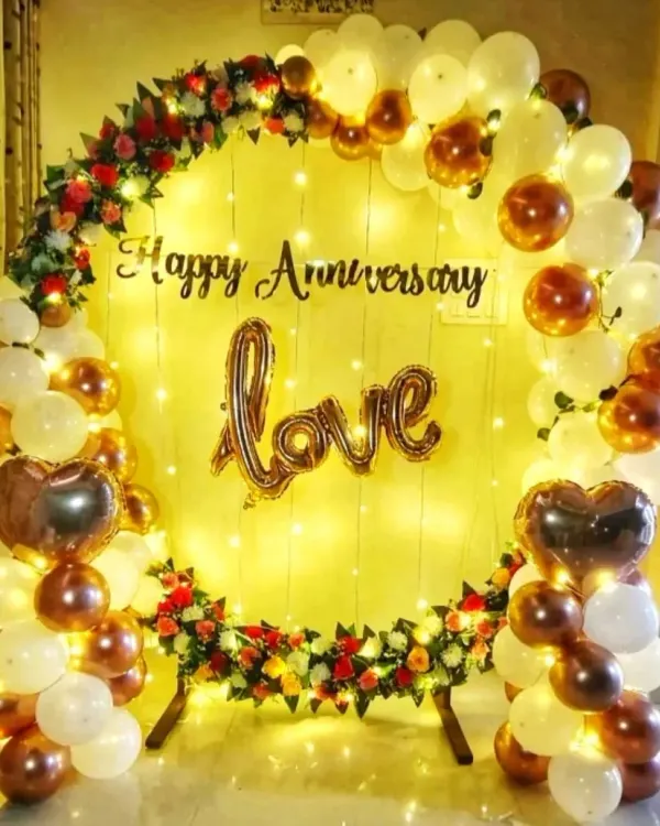 A romantic anniversary decoration for a home celebration. This setup features a circular backdrop with white and gold balloons, flowers, fairy lights, and a 'love' foil balloon.