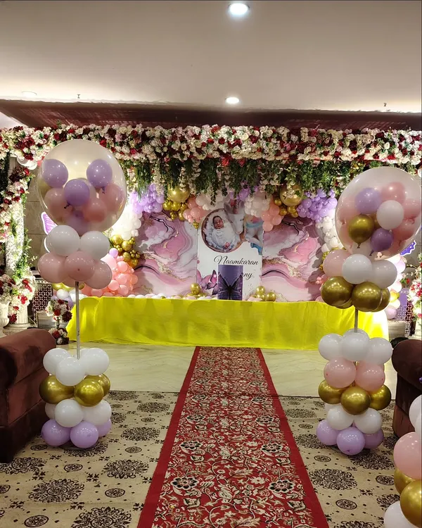 This is a lavish stage setup for a naming ceremony or first birthday. It includes a ceiling covered in artificial flowers, a custom backdrop, and elegant balloon pillars in pastel and gold tones.