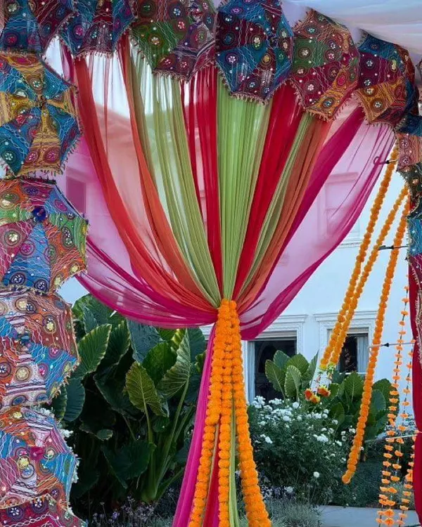 An outdoor garden setup for a Mehendi, featuring a canopy of colorful drapes and Rajasthani umbrellas. The decor is tied together with bright orange marigold garlands.