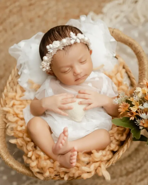A peaceful newborn girl, dressed in a delicate white outfit and floral headband, sleeping soundly in a cozy woven basket. We focus on creating a serene atmosphere for these early sessions.