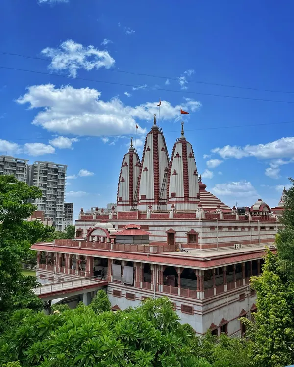 The beautiful white and red shikhara of the ISKCON NVCC temple in Pune, framed by lush green trees and a bright blue sky.