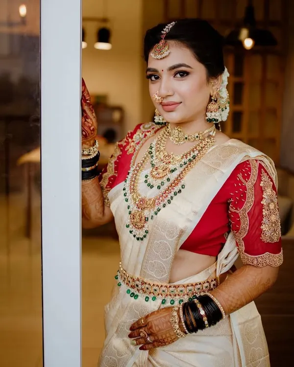 This is the quintessential South Indian bridal look I love to create. The layered antique necklaces, complete with a detailed choker, longer haar, maang tikka, and waist belt, come together to adorn this bride in her beautiful off-white and red Kanjeevaram saree.