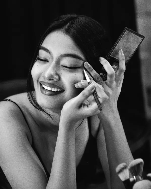 A candid, behind-the-scenes moment captured in timeless black and white. The monochrome treatment emphasizes the joy and emotion of the moment, focusing on the model's smile as she applies her makeup. It tells a story of preparation and passion.