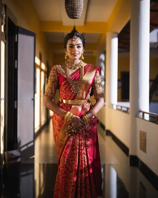 A stunning bride in a classic red Kanchipuram silk saree. The blouse features heavy gold embroidery, and the look is completed with traditional temple jewelry, perfect for a muhurtham ceremony.