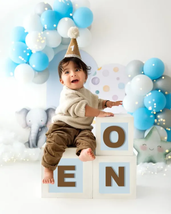 A one-year-old boy sits proudly on blocks that spell "ONE" during his first birthday photoshoot. The blue and white balloon arch adds a festive touch.