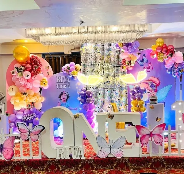 I create magical fairy and butterfly-themed birthday parties. This grand setup for a first birthday includes large character cutouts, custom marquee letters for the name, and a lush balloon arrangement in a party hall.