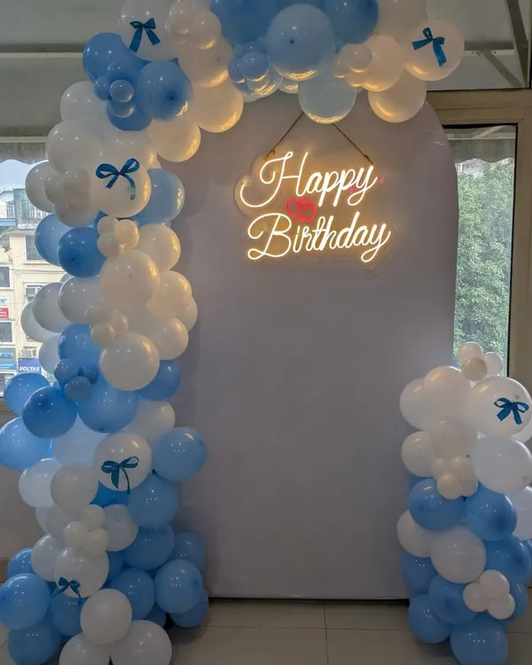 A beautiful balloon arch in pastel blue and white with a neon "Happy Birthday" sign, creating an elegant photo point or entrance for a party.
