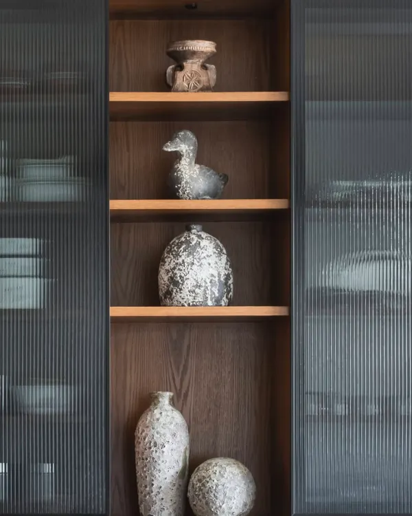 There’s a quiet poetry to rustic vases. Here, a collection of them is displayed in a fluted glass cabinet, their raw, weathered finish bringing depth and character to the polished space.