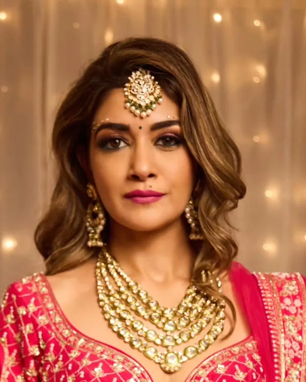 A glamorous bridal look featuring soft, side-swept waves and a dramatic smokey eye. The makeup is balanced with a neutral pink lip, allowing the intricate jewelry and vibrant pink lehenga to shine.