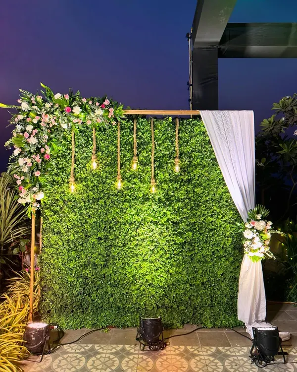 A modern and chic photo backdrop featuring a lush greenery wall. I added an asymmetrical floral arrangement, hanging Edison bulbs, and a simple white drape for a stylish and versatile photo corner.