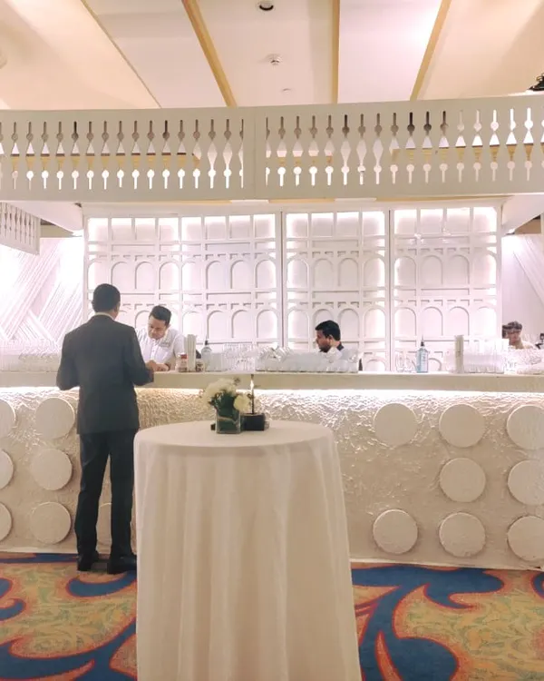 A stunning all-white bar setup for a high-end event. The design features intricate latticework, a textured front panel, and is flanked by large white floral arrangements, creating a sophisticated focal point for serving drinks.