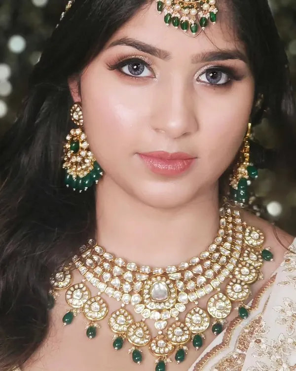 A beautiful close-up of a model wearing a classic Kundan necklace with green bead drops, paired with matching earrings and maang tikka.