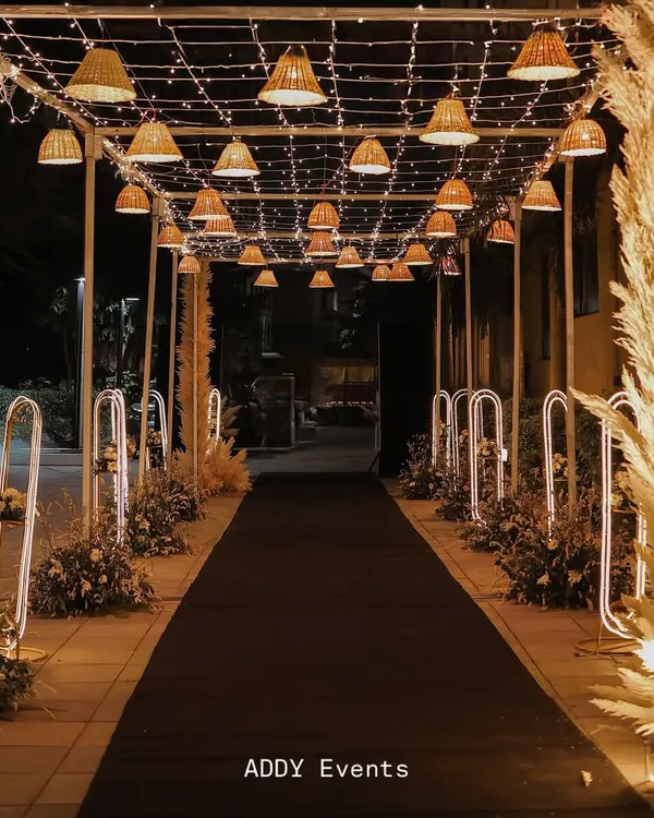 A magical walkway to an evening celebration. This enchanting passage is lit by a canopy of fairy lights and woven basket lamps, with the path flanked by glowing floral arrangements.