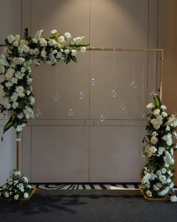 A minimalist and modern floral frame for a Bou-Bhaat or reception event. The asymmetrical arrangement of white flowers and hanging crystals creates a chic and elegant photo corner.