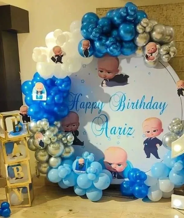 This Boss Baby themed decoration for a first birthday is one of my specialties. It features a custom printed circular backdrop, a large light-up number '1', transparent "BABY" display boxes, and a balloon garland in chrome blue, silver, and white.
