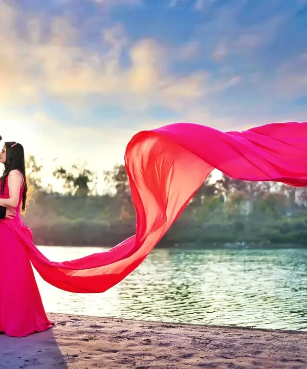 A breathtaking pre-wedding shot by the water, with a vibrant pink fabric catching the wind against the sunset sky. I use elements like flowing dresses to add drama and movement to my photographs.