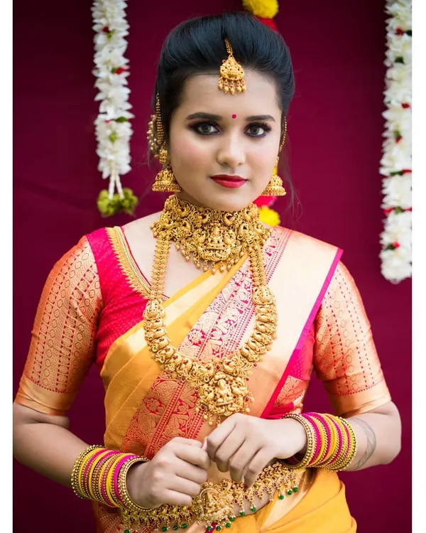 A classic muhurtham look featuring heavy temple jewelry and a bright yellow and red silk saree. The makeup is defined with bold eyes and a red lip to match the traditional attire.