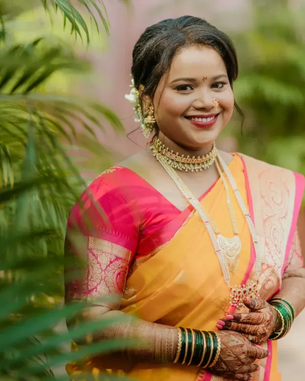 A radiant Maharashtrian bride, captured in natural light. Her look features a warm, glowing base, a classic red lip, and traditional gold jewelry, perfectly complementing her yellow and pink saree.