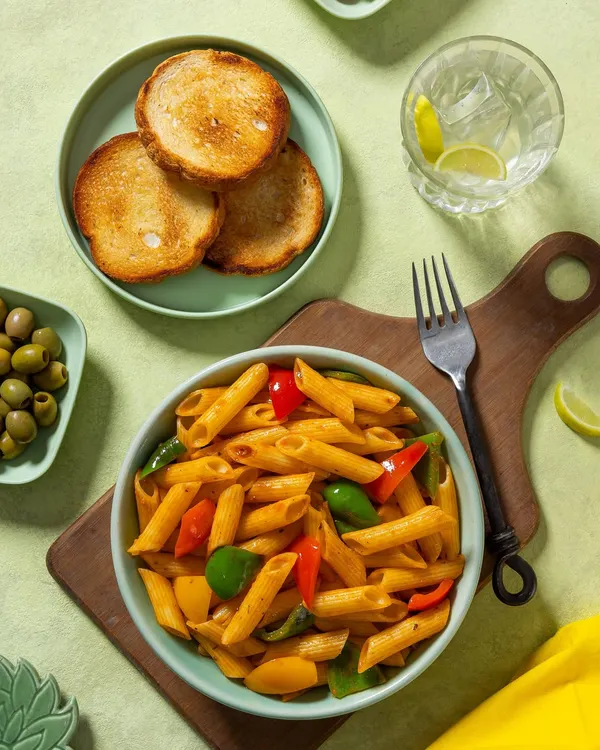 A bowl of penne pasta in a red sauce with bell peppers, shot for Rakesh Masala. The fresh, bright styling with a side of garlic bread makes for an inviting menu image.