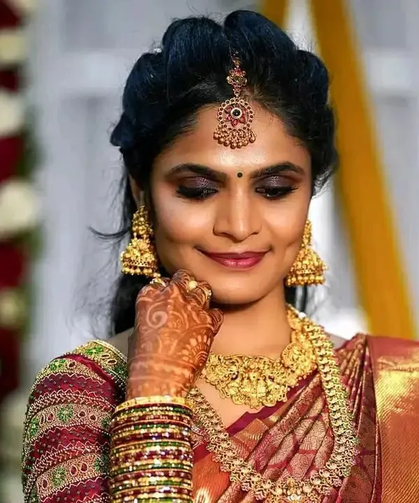 A detailed portrait of a bride's reception makeup. The look features classic gold jewelry, a neat hair updo with a maang tikka, and eye makeup that makes her features pop for the camera.