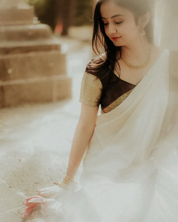 A serene portrait of the bride-to-be, captured in soft, natural light. Her traditional white and gold saree complements the classic stone architecture, creating a moment of quiet elegance.