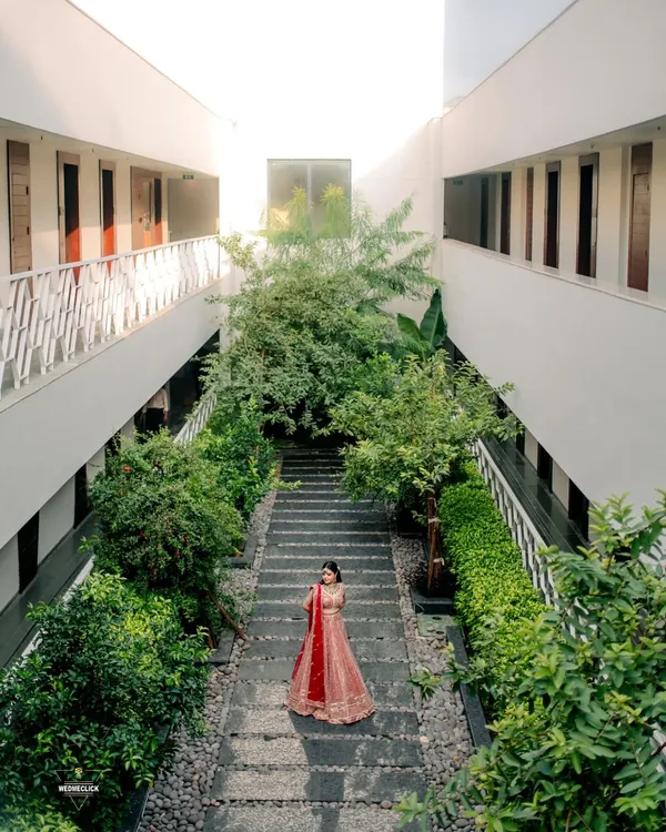 A stunning wide-angle shot of the bride in her red lehenga, framed by the modern architecture and lush greenery of the venue. This portrait showcases both the bride and her beautiful surroundings.