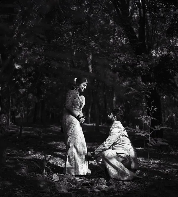 A dramatic, high-contrast black and white photo of a proposal in the forest. The monochrome edit emphasizes the light and shadow, creating a powerful and emotional scene.
