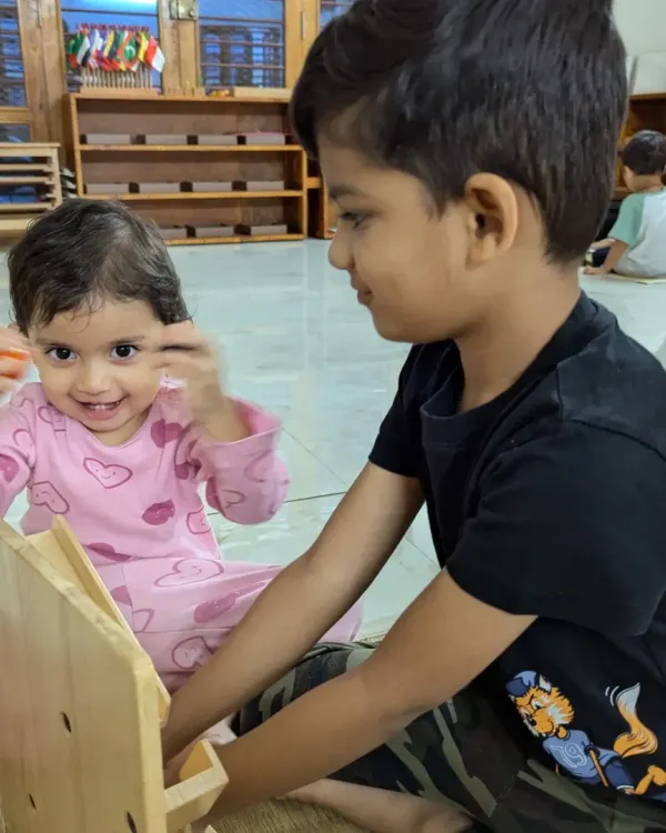 An older brother helps his younger sister with a wooden dressing frame, a practical life activity that develops fine motor skills. This kind of peer teaching happens naturally in our mixed-age group.