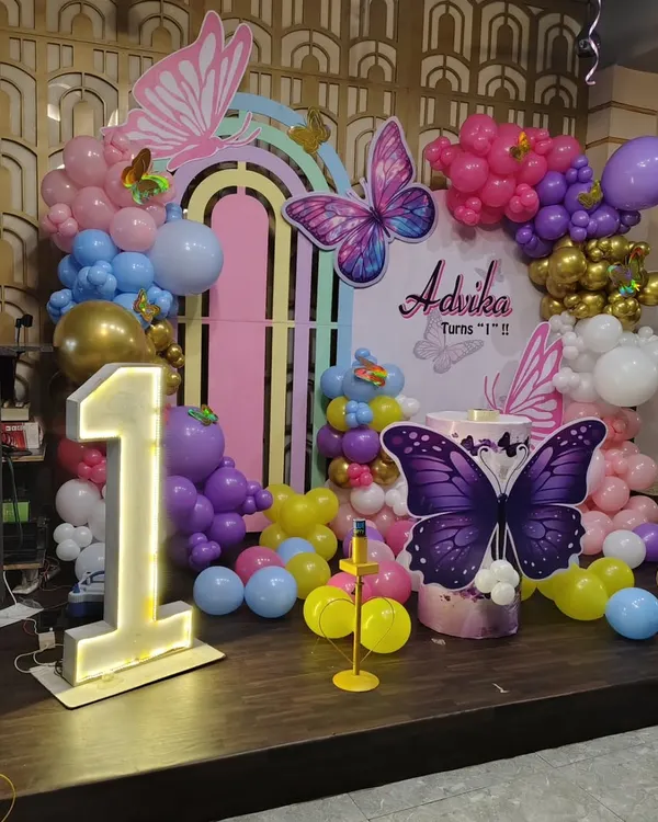 This is a butterfly-themed first birthday stage setup. It features a custom pastel rainbow backdrop, large butterfly cutouts, a light-up number '1', and a colorful balloon garland.