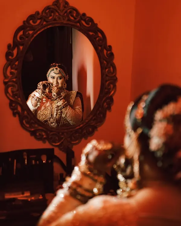 A creative bridal portrait using a mirror to capture the bride's reflection as she gets ready. The warm, reddish lighting adds a dramatic and intimate feel to this getting-ready shot.