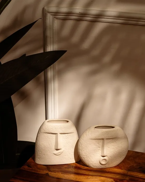 A pair of artistic, face-themed vases styled on a wooden table. The play of natural light and palm leaf shadows creates a warm, serene, and stylish atmosphere.