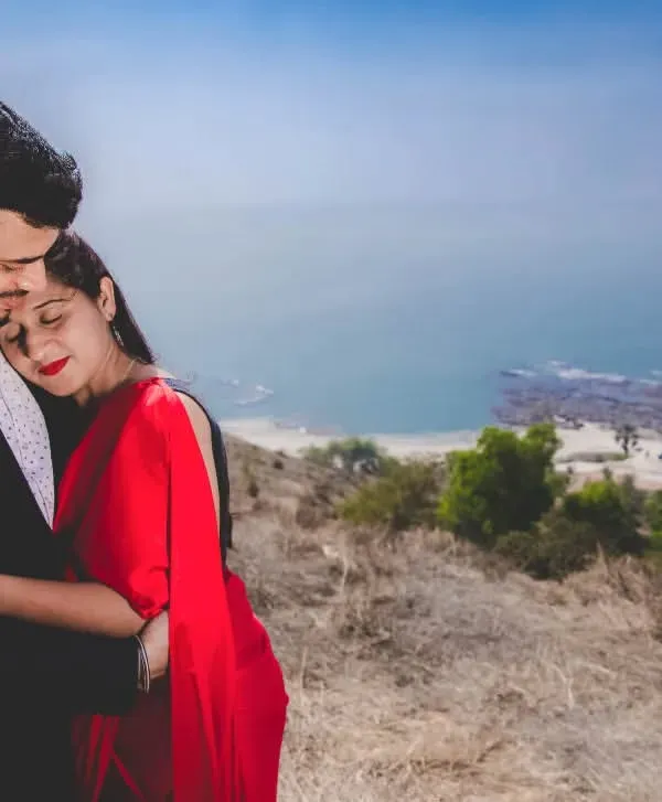 A tender embrace on a hillside overlooking the ocean. The woman's vibrant red dress stands out against the natural landscape, creating a passionate and romantic pre-wedding portrait.