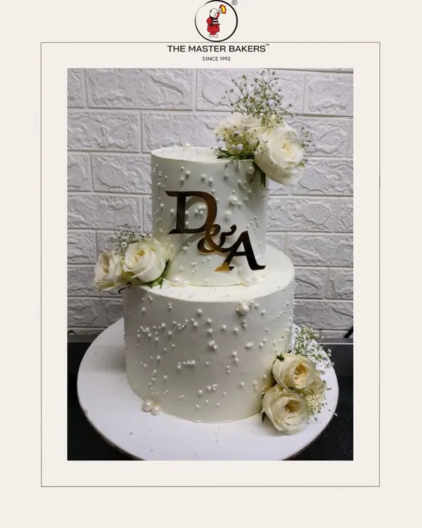 This two-tiered white engagement cake is a perfect example of timeless elegance. Adorned with delicate sugar pearls, fresh white roses, and a custom monogram, it’s designed to be a stunning centerpiece for your special day.