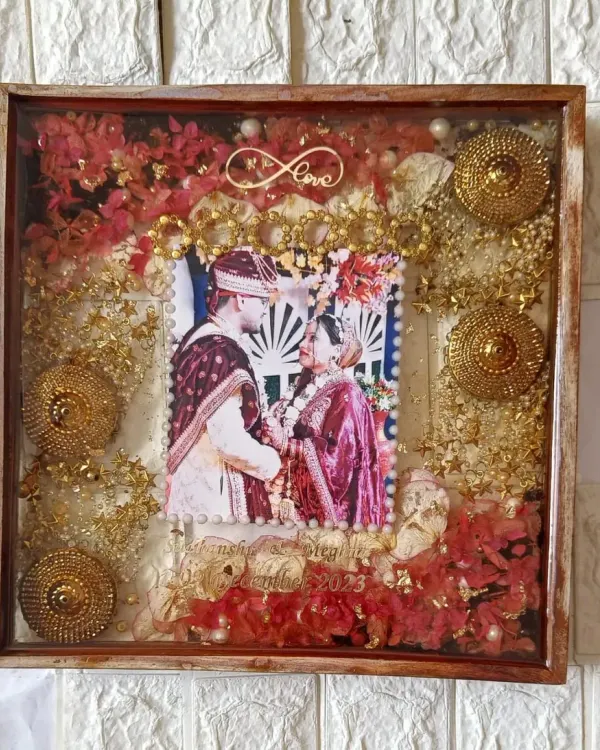 This square wedding keepsake frame is filled with vibrant red flowers and golden ornaments from the ceremony. The couple's photo is centered, surrounded by memories of their special day, including an infinity love symbol.