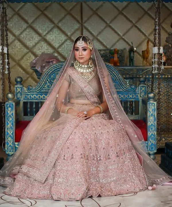 A wide shot of a bride in a magnificent pink lehenga, seated on a traditional blue swing. This composition captures the full grandeur of the bridal ensemble and the ornate setting.