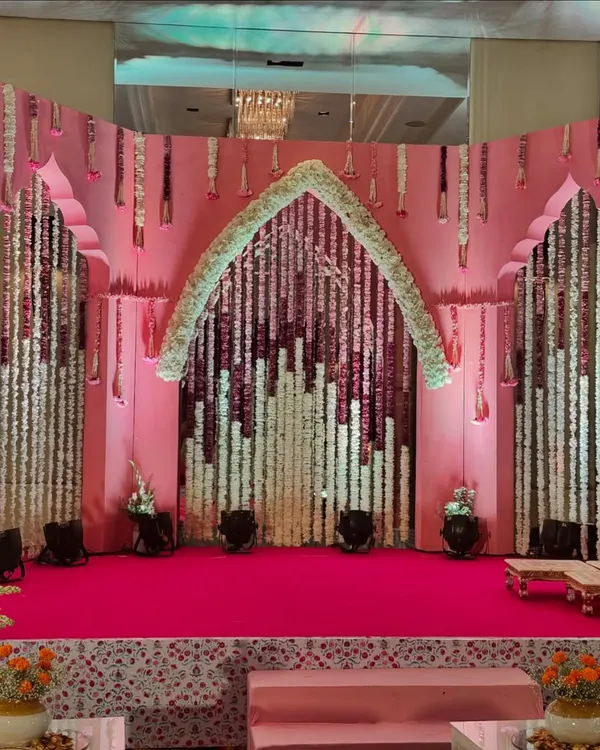 The stage for a 'Bhaat & Tilak' ceremony. The decor uses a blend of traditional elements like floral strings and modern aesthetics with a pink and white color scheme.
