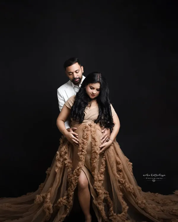 This moody portrait uses a dark backdrop to create a dramatic and intimate feel. The focus is entirely on the couple and the beautiful silhouette of the baby bump, highlighted by the stunning ruffled gown.