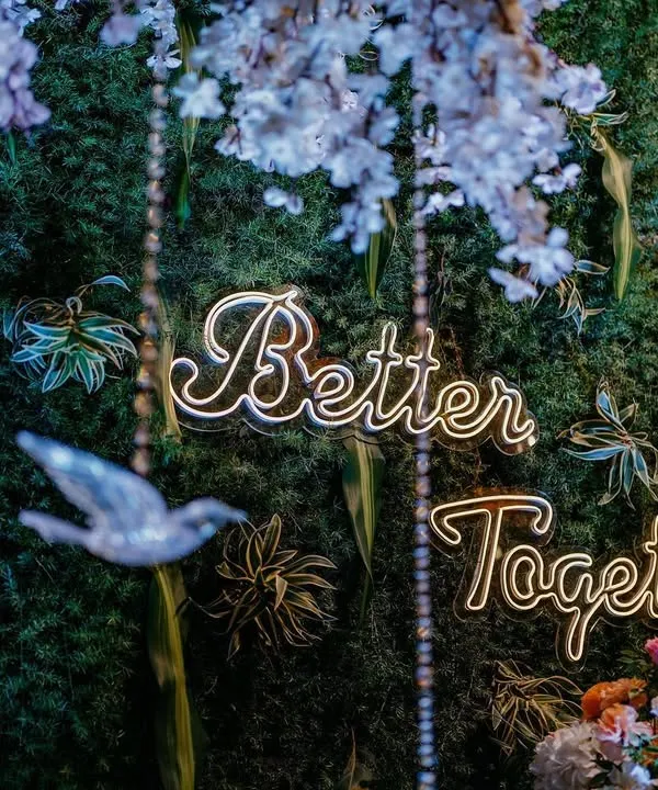 Love is in every detail. This close-up shows our "Better Together" neon sign set against a lush foliage wall, with delicate crystal birds hanging from cherry blossom branches.