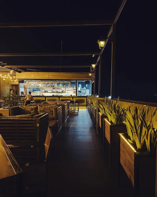 An atmospheric shot of a rooftop cafe at night. The design features an open view into the brightly lit kitchen, slatted wood seating, and a row of illuminated planters creating a pathway.
