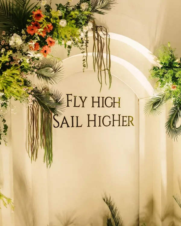 For a joint 50th and 80th birthday, we designed this elegant arched backdrop with the message "Fly High Sail Higher". Framed with lush tropical greenery and florals, it served as an inspiring focal point for the celebration.
