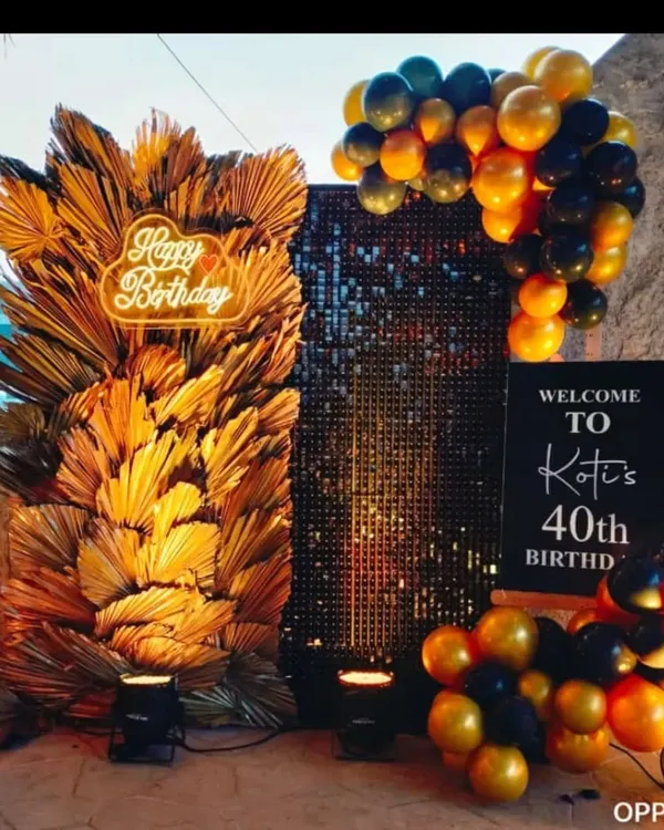 A sophisticated 40th birthday photo booth. The decor features a black sequin wall, a large arrangement of dried golden palm leaves, and a custom neon sign.