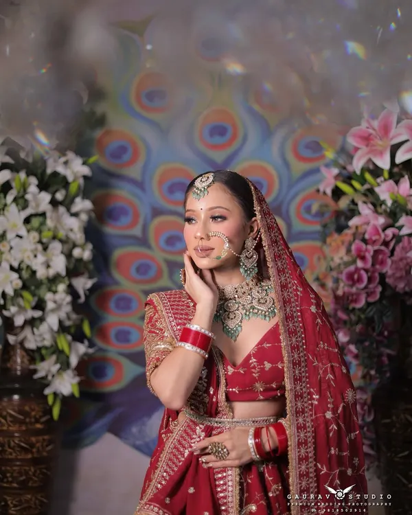 A mid-shot of the bride in a red embroidered lehenga, set against a floral and peacock-themed backdrop.