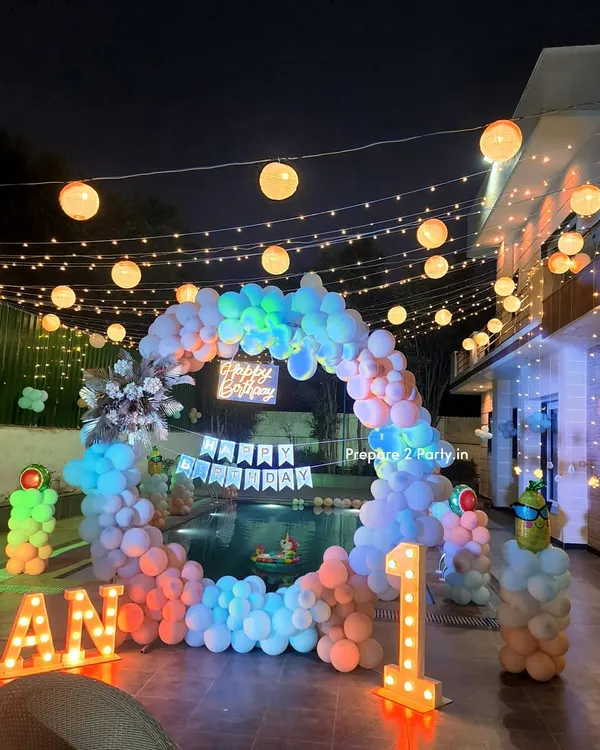 This stunning poolside first birthday setup is perfect for a grand celebration. The circular balloon arch frames the water beautifully, while marquee letters and extensive string lighting create a magical, festive atmosphere for an evening party.