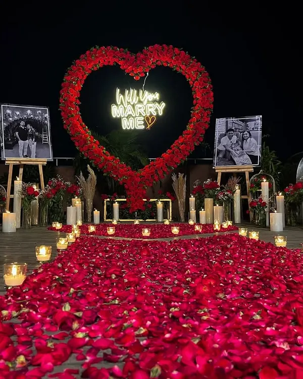 A stunning outdoor marriage proposal setup on a terrace. We created this unforgettable moment with a large heart-shaped arch made of red florals, a custom 'Will you MARRY ME' neon sign, and a romantic walkway of rose petals and candles.