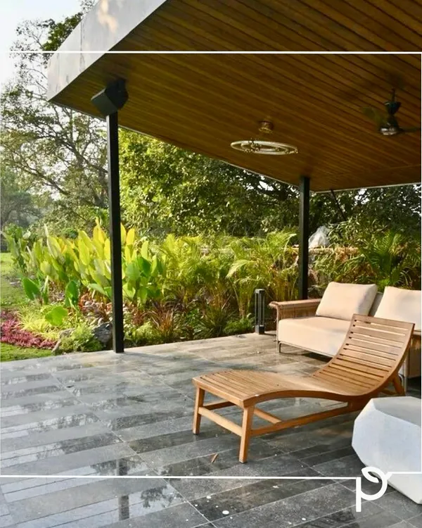 A covered patio overlooking a lush garden we designed. The space features comfortable wooden lounge furniture and is bordered by a dense bed of tropical plants, creating a perfect blend of indoor comfort and outdoor nature.