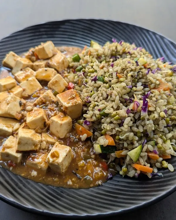 This is a plant-based protein meal featuring tofu in a savory garlic pepper sauce, served alongside pesto-flavored brown rice mixed with vegetables. It's a flavorful and complete vegetarian meal.